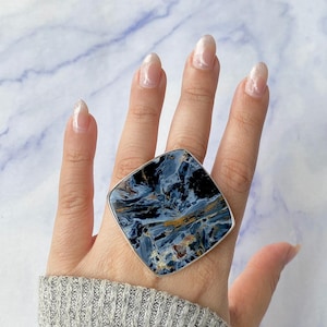 Może przedstawiać: Duży, kwadratowy pierścionek z wypolerowanym niebiesko-czarnym kamieniem szlachetnym ze złotymi i brązowymi żyłkami, osadzony w srebrnym metalu. Pierścionek jest noszony na dłoni, prezentując skomplikowane wzory i kolory kamienia.