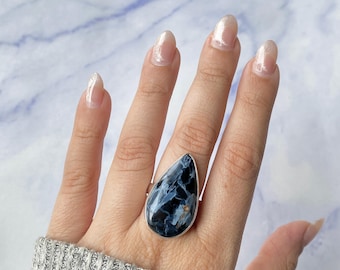 May include: A teardrop-shaped ring featuring a polished blue and black patterned stone set in a silver-toned metal band. The stone displays swirling dark navy, charcoal, and light blue veins throughout its surface, secured by a smooth metal bezel.