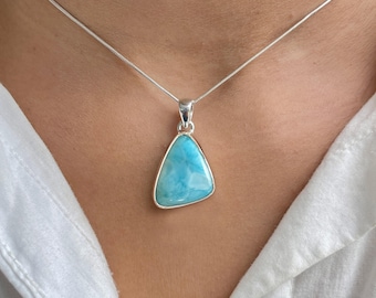 May include: A silver necklace featuring a triangular pendant with a polished, light blue Larimar stone. The stone displays natural white marbling patterns and is set in a smooth silver bezel. The pendant hangs from a thin, flexible silver chain.