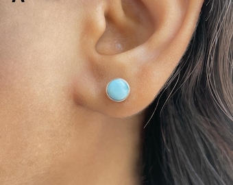 May include: A round stud earring featuring a smooth, light blue cabochon stone set in a polished silver-toned metal bezel. The earring is positioned in the earlobe, showcasing a simple, circular design with a clean, reflective metallic edge.