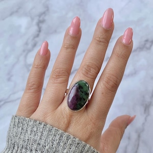 Pode incluir: Um anel de prata com uma grande gema oval verde e rosa. O anel está em um dedo.