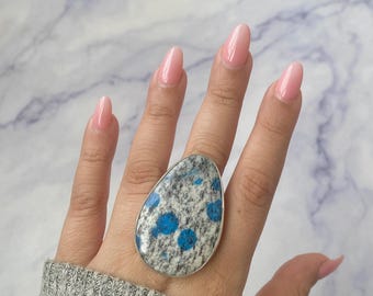May include: A large teardrop-shaped ring featuring a speckled gray and white stone with vibrant blue circular inclusions. The stone is set in a simple silver-toned metal bezel, creating a bold statement piece that covers a significant portion of the finger.