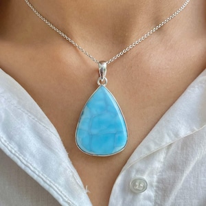 以下が含まれることがあります： 銀色のネックレスで、涙型のペンダントが付いています。ペンダントは、銀色のベゼルにセットされた、薄い青色のマーブル模様です。白い襟付きのシャツの上にネックレスが着用されています。