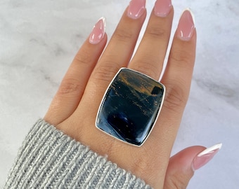 May include: A large rectangular ring featuring a polished dark blue and gold tiger eye gemstone set in a smooth silver metal bezel. The stone displays deep navy tones with shimmering golden streaks across its surface, mounted on a simple band.