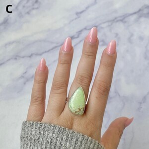 May include: A teardrop-shaped light green stone ring set in a silver-toned metal bezel. The stone features natural brown matrix inclusions throughout its surface. The ring is marked with the letter C in the upper left corner of the frame.
