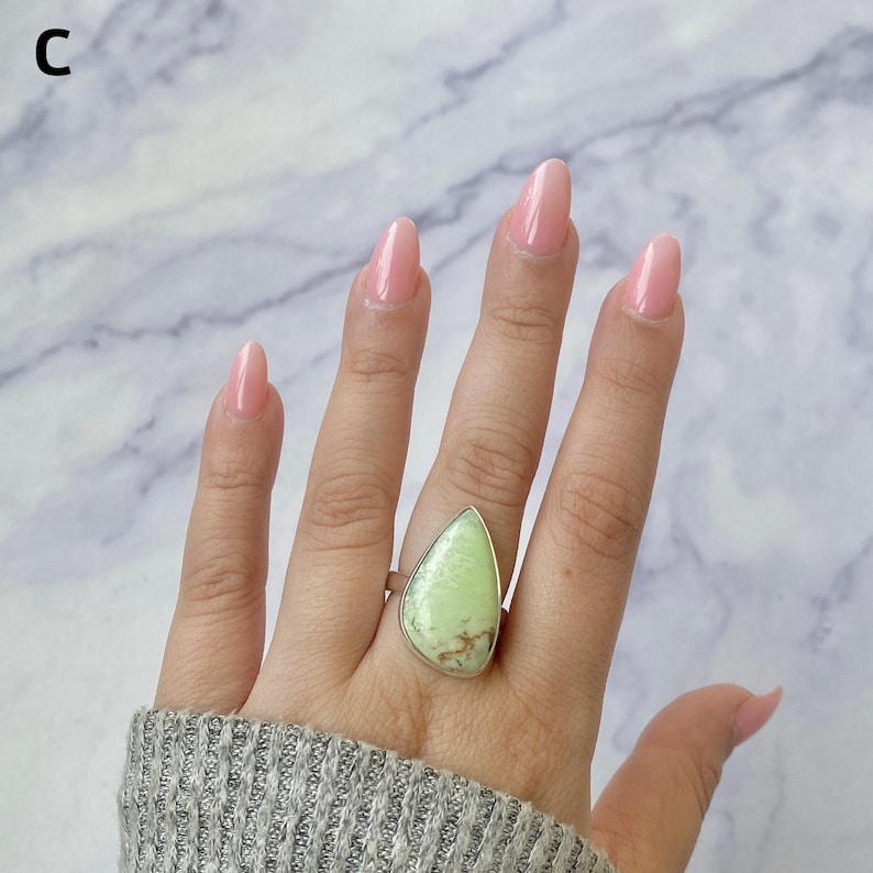 May include: A teardrop-shaped light green stone ring set in a silver-toned metal bezel. The stone features natural brown matrix inclusions throughout its surface. The ring is marked with the letter C in the upper left corner of the frame.