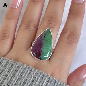 May include: A silver ring featuring a large, teardrop-shaped cabochon stone with a distinct split color pattern. One half of the polished gemstone is a deep magenta purple, while the other half is a vibrant forest green, set in a smooth metal bezel.