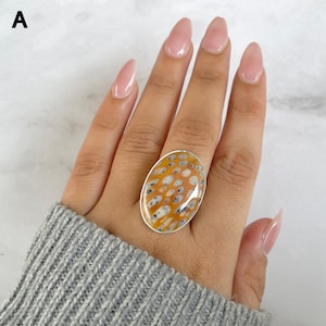 May include: An oval-shaped ring featuring a polished stone with a vibrant orange background and a pattern of small, dark, irregular spots. The stone is set in a simple silver-toned metal bezel, creating a smooth, rounded profile on the finger.