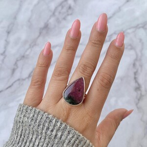 Peut inclure: Bague en argent ornée d'une grosse pierre en forme de goutte d'eau. La pierre présente des teintes rouge foncé et vertes. La bague est portée sur une main vêtue d'un pull gris, avec du vernis à ongles rose.