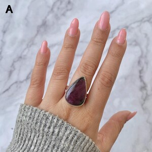 May include: A silver ring featuring a large, teardrop-shaped, deep purple gemstone with visible dark inclusions. The stone is set in a smooth metal bezel, and the band is made of polished silver. The letter A is visible in the top left corner.