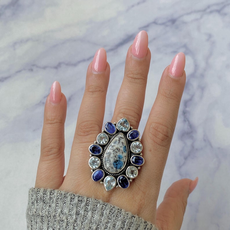 K&ouml;nnte beinhalten: Ein silberner Ring mit einem gro&szlig;en blauen und wei&szlig;en Edelstein, umgeben von kleineren blauen und wei&szlig;en Edelsteinen.