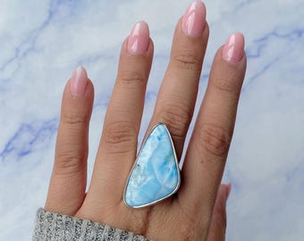 May include: A large, triangular-shaped Larimar gemstone ring set in a smooth silver bezel. The stone features a marbled pattern of sky blue and white tones with a polished, glossy surface. The ring is worn on a finger, showcasing the stone's size.