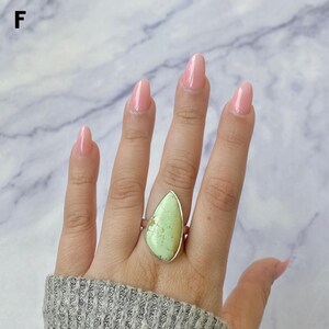 May include: A ring featuring a large, pear-shaped, light green stone with subtle darker green inclusions, set in a simple metal band. The stone has a smooth, polished surface and is positioned centrally on the finger, showcasing its unique shape.