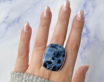May include: A large statement ring featuring a rectangular, polished blue and black patterned stone set in a simple silver-toned metal band. The stone displays a complex, marbled texture with deep navy and lighter sky blue veins throughout the surface.