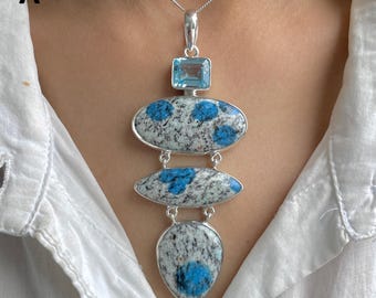 Könnte beinhalten: Eine silberne Anhängerkette mit einem quadratischen hellblauen Edelstein an der Spitze, gefolgt von drei vertikal verbundenen Steinen mit einem gesprenkelten grau-weißen Muster und leuchtend blauen kreisförmigen Einschlüssen, an einer dünnen Silberkette.