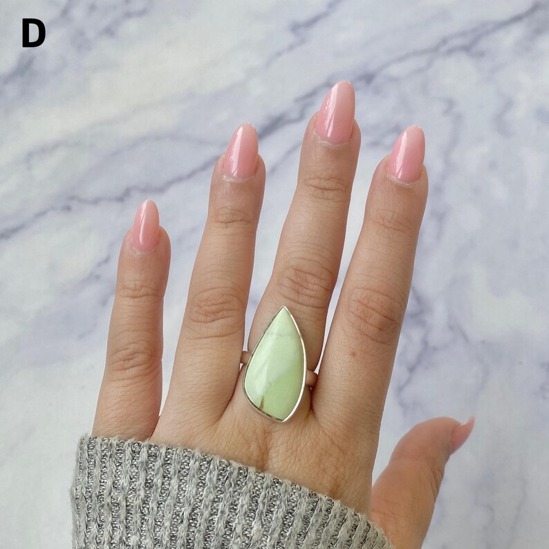May include: A silver ring featuring a large, teardrop-shaped, light green stone set in a smooth metal bezel. The stone displays subtle natural veining and a matte finish. The letter D is visible in the upper left corner of the image.