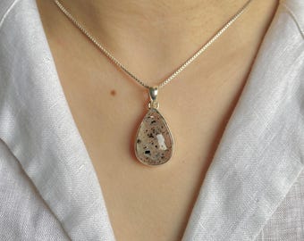 Op de afbeelding: Een zilveren ketting met een druppelvormige hanger van een gladde, doorschijnende steen met kleine zwarte spikkels. De hanger zit vast aan een eenvoudig zilveren oogje en hangt aan een fijne, dunne zilveren ketting met vierkante schakels.