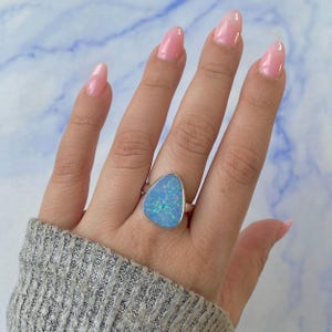 Può includere: Anello in argento con una grande pietra di opale blu triangolare. L'opale presenta riflessi iridescenti verdi e blu. L'anello è posto su uno sfondo azzurro e bianco.