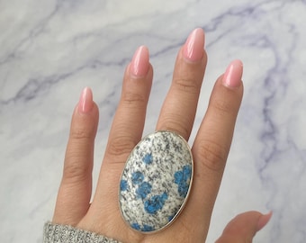 May include: A large oval ring featuring a speckled gray and white stone with prominent blue inclusions. The stone is set in a simple silver-toned metal bezel, creating a bold statement piece that covers a significant portion of the finger.