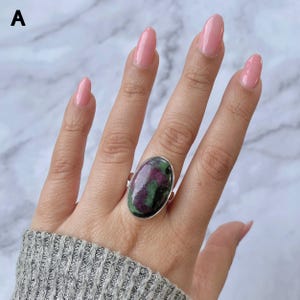 Könnte beinhalten: Ein Silberring mit einem großen, polierten, ovalen Cabochon-Stein, der ein gesprenkeltes Muster aus tiefem Magenta, Dunkelgrün und schwarzen Einschlüssen aufweist. Der Stein ist in einer schlichten, glatten Silberfassung eingefasst.