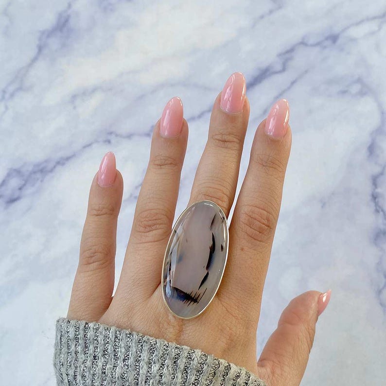 May include: A silver ring with a large oval-shaped agate stone. The stone is a mix of white, gray, and black colors. The ring is on a person's finger.