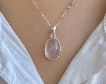 Op de afbeelding: Een zilveren ketting met een fijne figaro-schakelketting en een ovaalvormige hanger. De hanger bevat een doorschijnende, lichtgekleurde steen met zichtbare interne insluitingen, gevat in een gladde, gepolijste zilveren zetting.