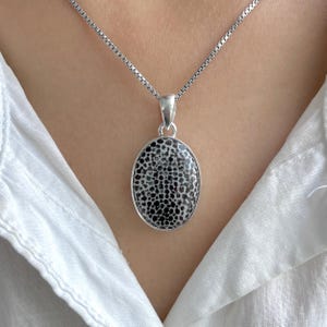May include: Silver pendant necklace featuring an oval-shaped, black and white patterned stone. The pendant hangs from a delicate silver chain. The necklace is displayed against a white shirt.