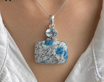 Könnte beinhalten: Ein rechteckiger Anhänger mit einem gesprenkelten weiß-schwarzen Stein mit leuchtend blauen Einschlüssen, gekrönt von einem kleinen ovalen, facettierten hellblauen Edelstein. Der Anhänger ist in einen silbernen Rahmen gefasst und an einer Kette.