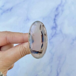 May include: A silver ring with a large oval-shaped dendritic agate stone. The stone is a light gray color with brown and black dendritic patterns.