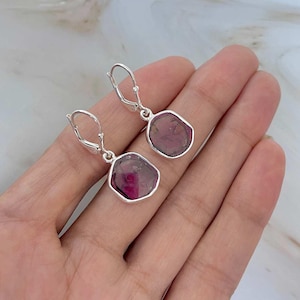 May include: A pair of silver leverback earrings. Each earring features a faceted, irregular-shaped, dark pink gemstone set in a silver bezel. The gemstones have a translucent quality, with variations in color. The earrings are displayed against a neutral background.