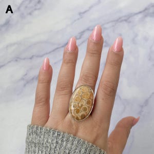 Pode incluir: Um anel com um grande cabochão oval de coral fossilizado com um padrão de favo de mel em tons de castanho e bege. A pedra está engastada numa moldura simples de metal prateado polido que contorna o perímetro da pedra fóssil oval.
