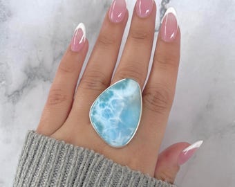 May include: A large, irregular, teardrop-shaped larimar gemstone ring set in a smooth silver bezel. The stone features a marbled, sky-blue and white pattern reminiscent of ocean waves. The ring is worn on a finger, showcasing the stone's polished surface.