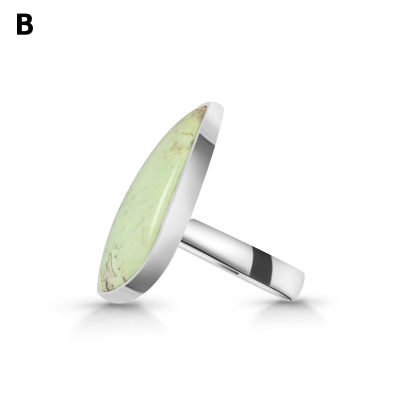 May include: A silver ring featuring a large, elongated, teardrop-shaped stone in a pale mint green colour with natural tan veining. The metal band is smooth and polished, showcasing a clean, metallic finish that contrasts with the textured stone.