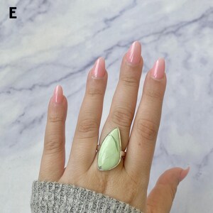 May include: A ring featuring a large, teardrop-shaped, light green stone with subtle dark veining, set in a simple silver-toned metal band. The stone has a smooth, polished surface and is positioned vertically on the finger.