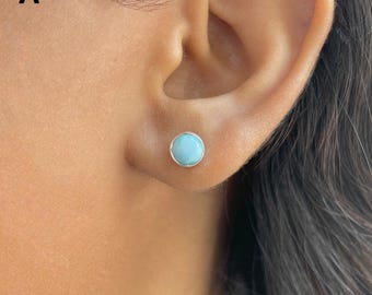 May include: A round stud earring featuring a smooth, light blue stone set in a polished silver-coloured metal bezel. The circular gemstone has a solid, opaque appearance and is centred on the earlobe, showcasing a simple and clean design.