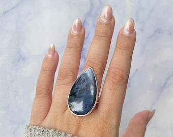 May include: A teardrop-shaped blue kyanite gemstone ring set in a simple silver bezel. The stone features deep blue and dark grey streaks with a polished, reflective surface. The ring is worn on a finger, showcasing the large, prominent stone.