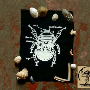 Peut inclure: Un patch en tissu noir avec une illustration blanche d'un scarabée avec des parties du corps étiquetées, y compris la tête, le thorax, l'abdomen et la patte. Le texte "georupidae" est également inclus. Le patch est entouré de coquillages.