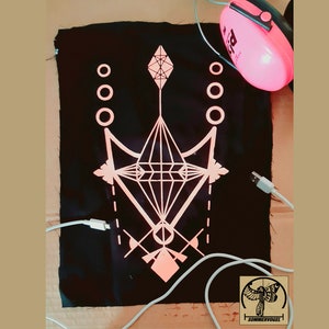 Peut inclure: Un patch en tissu noir avec un motif géométrique rose. Le motif présente une forme de diamant avec des diamants plus petits et des cercles autour. Le patch est entouré de cordons blancs.