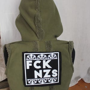 Könnte beinhalten: Olivgrüne Kapuzenweste mit einem schwarz-weißen Aufnäher mit der Aufschrift "FCK NZS". Die Weste hat einen flauschigen Besatz an Kapuze und Kanten. Die Kapuze ist hochgezogen, und die Weste wird auf einer Schaufensterpuppe präsentiert.