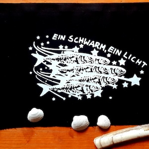 Könnte beinhalten: Schwarzer Stoff mit einem weißen Design aus Fischskeletten und Sternen, mit dem Text "EIN SCHWARM, EIN LICHT". Das Bild ist von Muscheln und anderen natürlichen Objekten umgeben, was ein Küstenmotiv erzeugt.