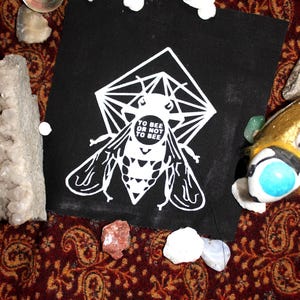 Könnte beinhalten: Schwarzer Stoff-Patch mit einem weißen geometrischen Design, das eine Biene mit dem Text "To Bee Or Not To Bee" zeigt.