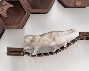 May include: A cat walks across a rope bridge attached to a wall-mounted cat tree made of dark wood. The cat tree has multiple hexagonal shelves and a curved platform.