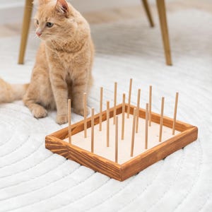 Könnte beinhalten: Ein Katzenspielzeug aus Holz mit einem rechteckigen Sockel und einem Gitter aus Holzstiften. Eine rotbraune Katze beobachtet das Spielzeug.