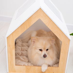 May include: A house-shaped pet bed featuring a light-colored wooden frame and a white roof with ventilation slats. Inside the structure, a fluffy, cream-colored dog with long fur rests on a soft, quilted beige cushion that lines the floor.
