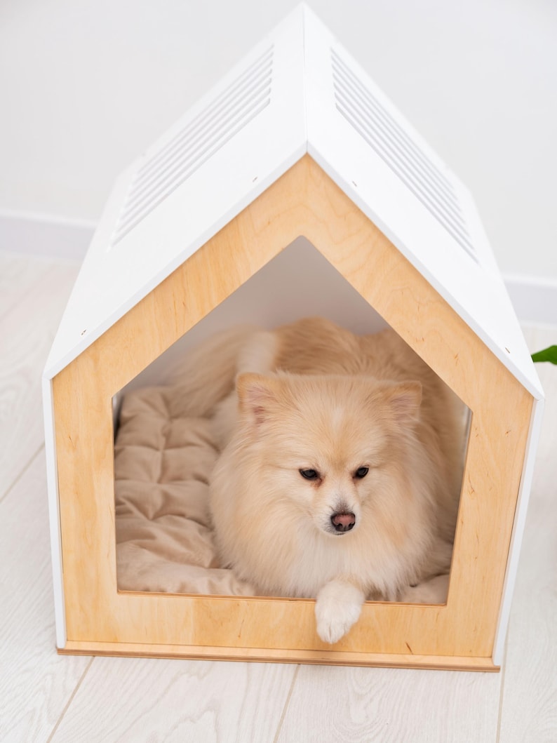 May include: A house-shaped pet bed featuring a light-colored wooden frame and a white roof with ventilation slats. Inside the structure, a fluffy, cream-colored dog with long fur rests on a soft, quilted beige cushion that lines the floor.