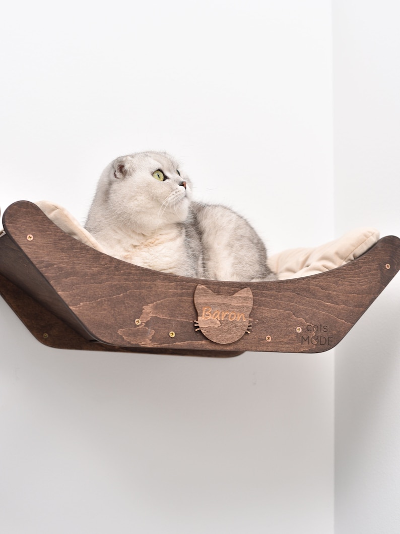 May include: A white cat with green eyes sits in a wooden cat bed. The bed is mounted on a wall and has a cat-shaped cutout with the name "Baron" inscribed on it. The bed is made by "Cats Mode".