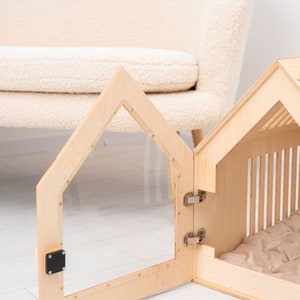 May include: A wooden dog house with a clear plastic door and a beige cushion inside. The door is open and the house is sitting on a light wood floor.