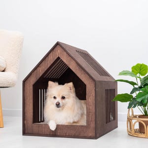 May include: A dark brown wooden pet house with a gabled roof and vertical slat cutouts on the sides. A small, cream-colored fluffy dog with pointed ears is resting inside the structure, looking forward with its front paw resting on the edge.