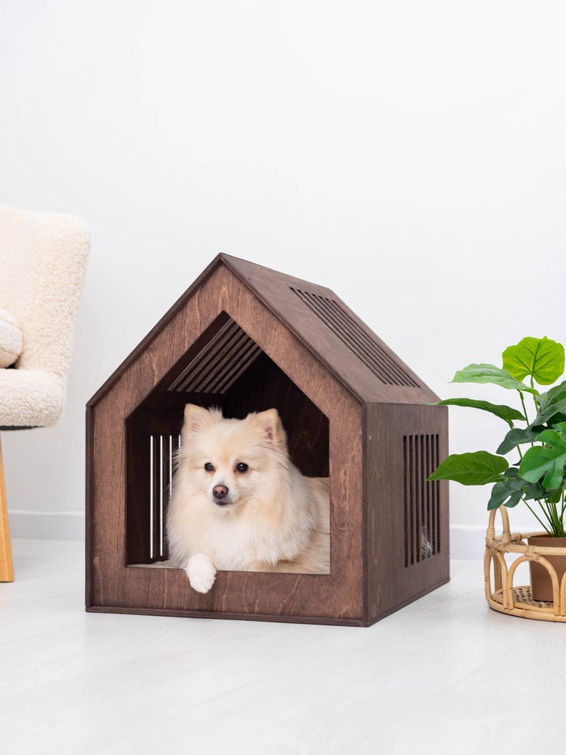 May include: A dark brown wooden pet house with a gabled roof and vertical slat cutouts on the sides. A small, cream-colored fluffy dog with pointed ears is resting inside the structure, looking forward with its front paw resting on the edge.