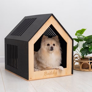 May include: A house-shaped pet bed with black exterior walls and a natural light wood front panel. The front panel features the name Buddy engraved in a script font. A small, cream-colored, long-haired dog is resting inside the structure.